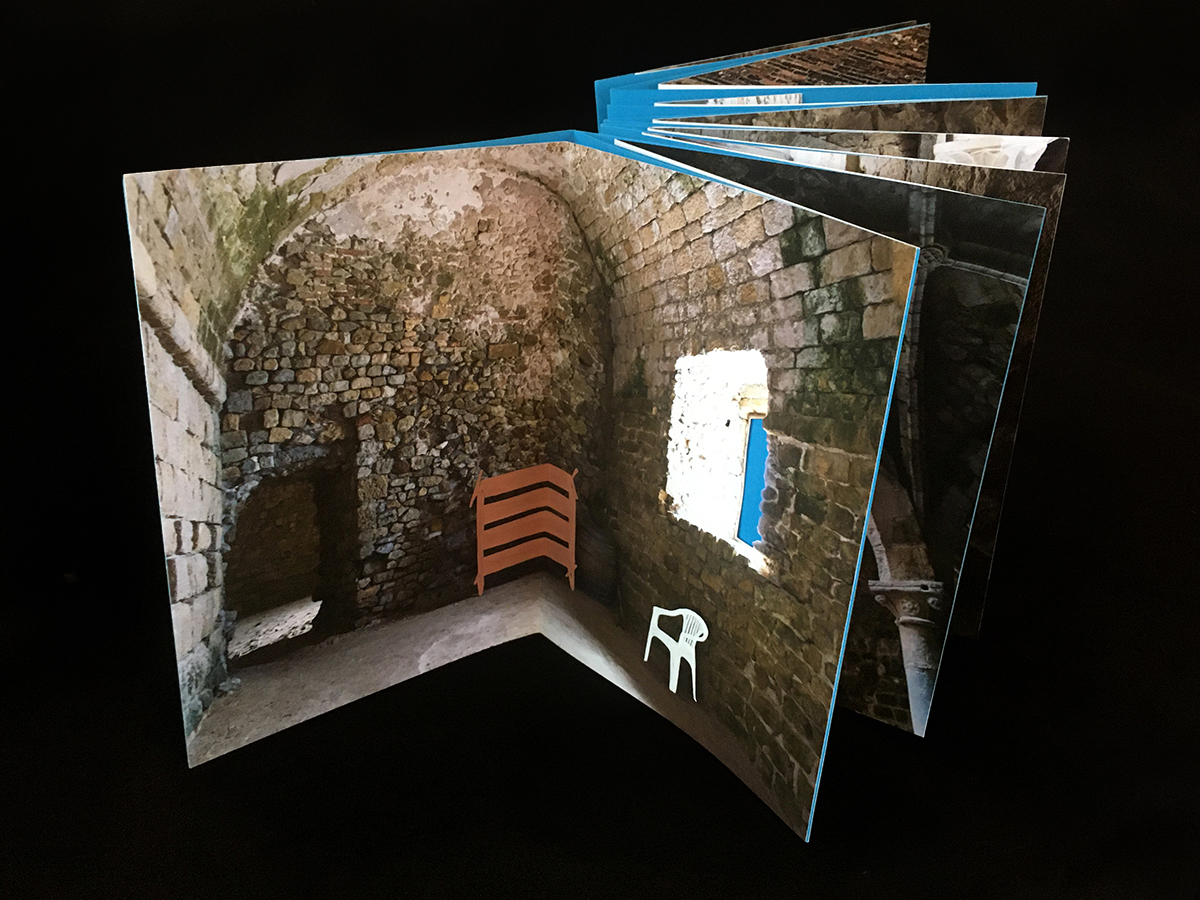 open page of the book show a photograph of an abandoned stone room with the window cut out and a plastic chair and shelf painted on