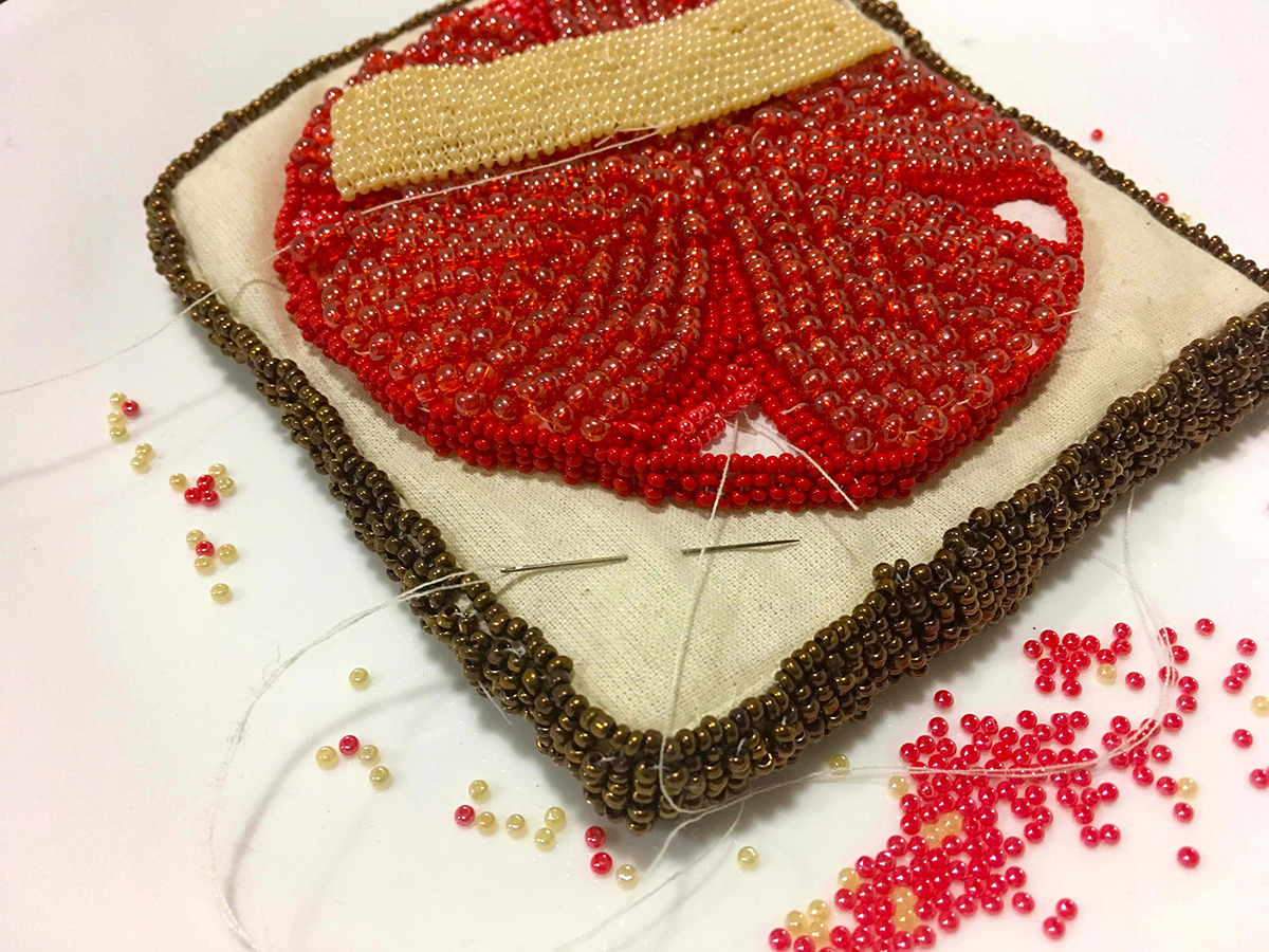 sculpture of an open face tomato and cheese sandwich made from fabric and glass beads. Thread, needle and extra beads show that the construction is in progress  