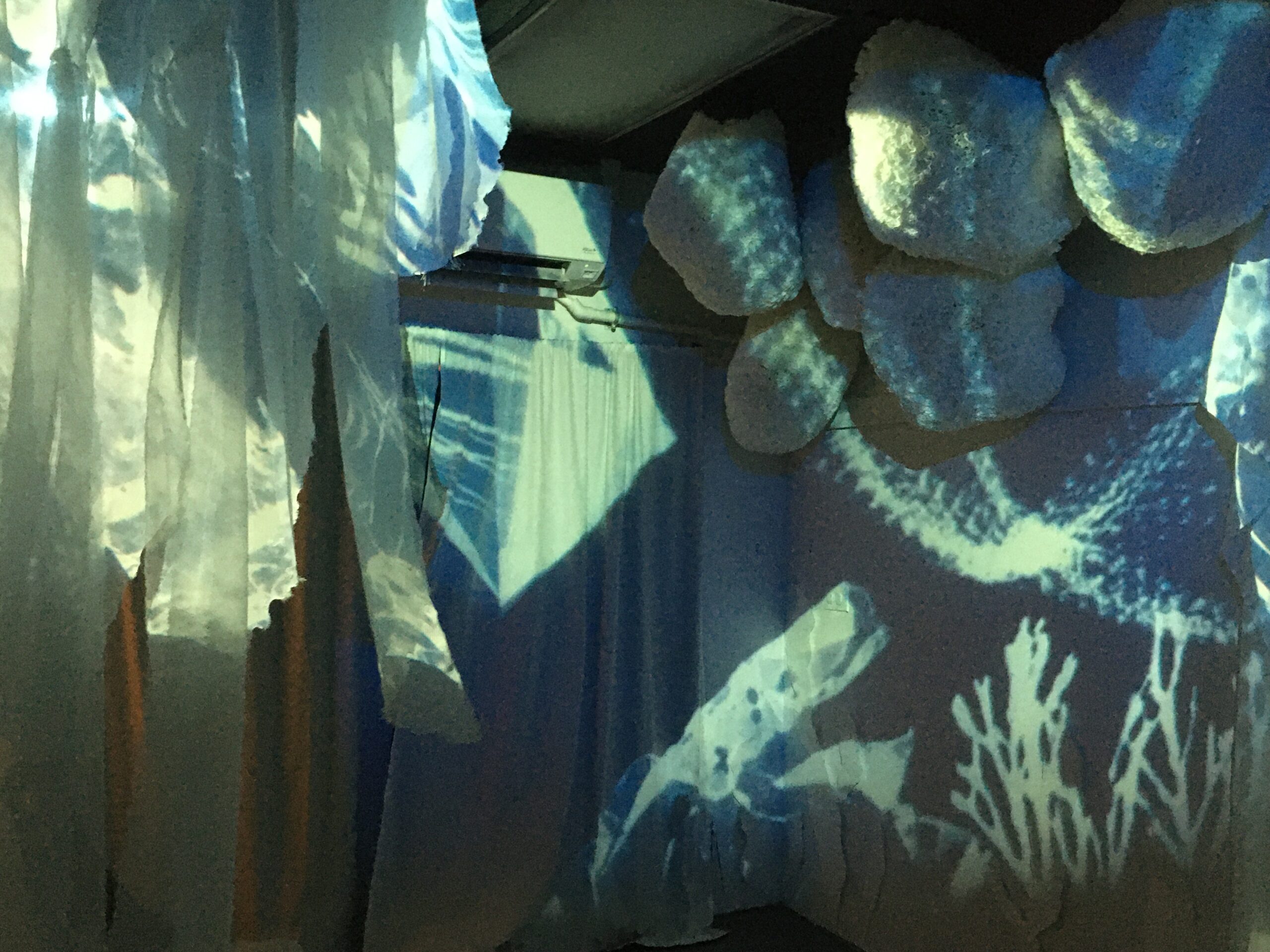 room filled with projection of cyanotype print of seaweed and plastic trash. Paper orb sculptures and translucent fabric hangs from the ceiling. 