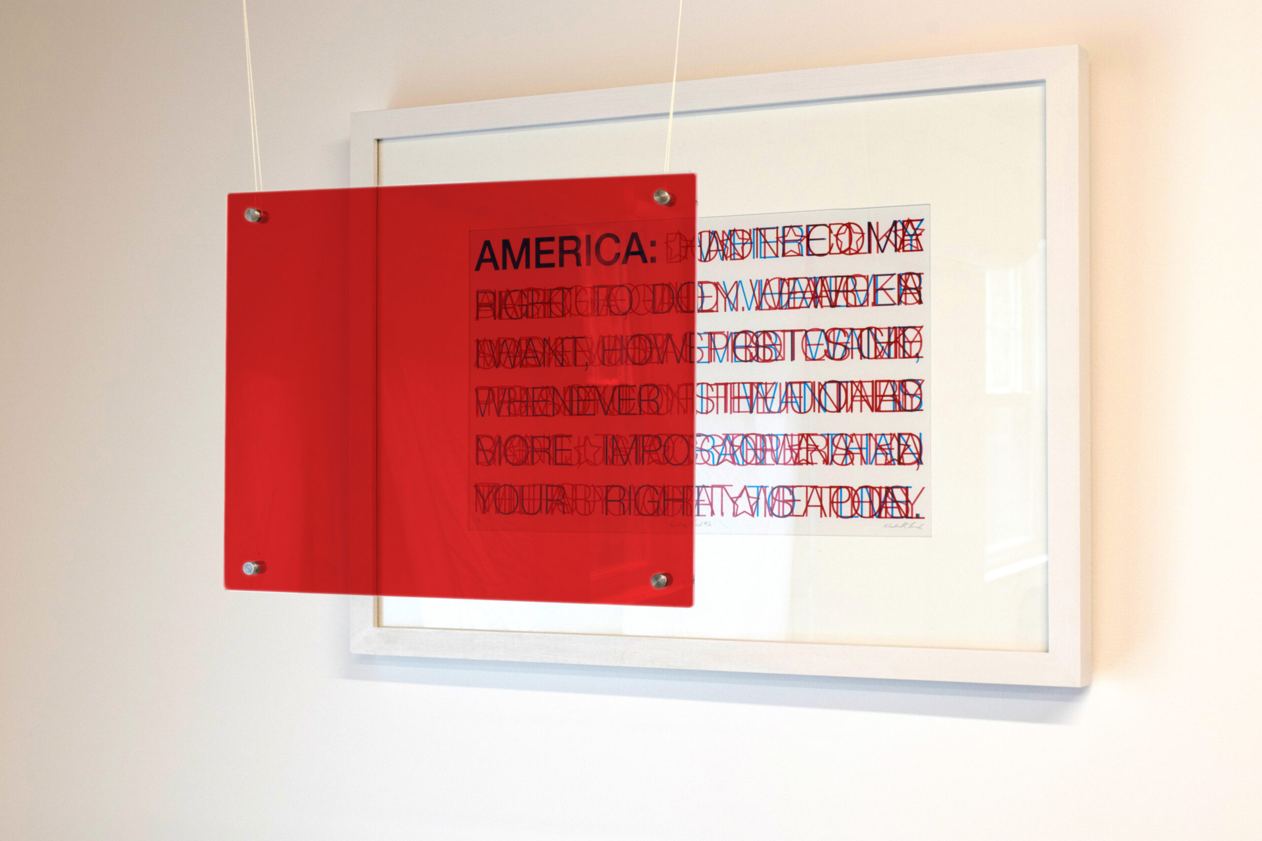 a framed screenprint of layers of red and blue text reminiscent of an American flag design with a red acrylic sheet hanging in front