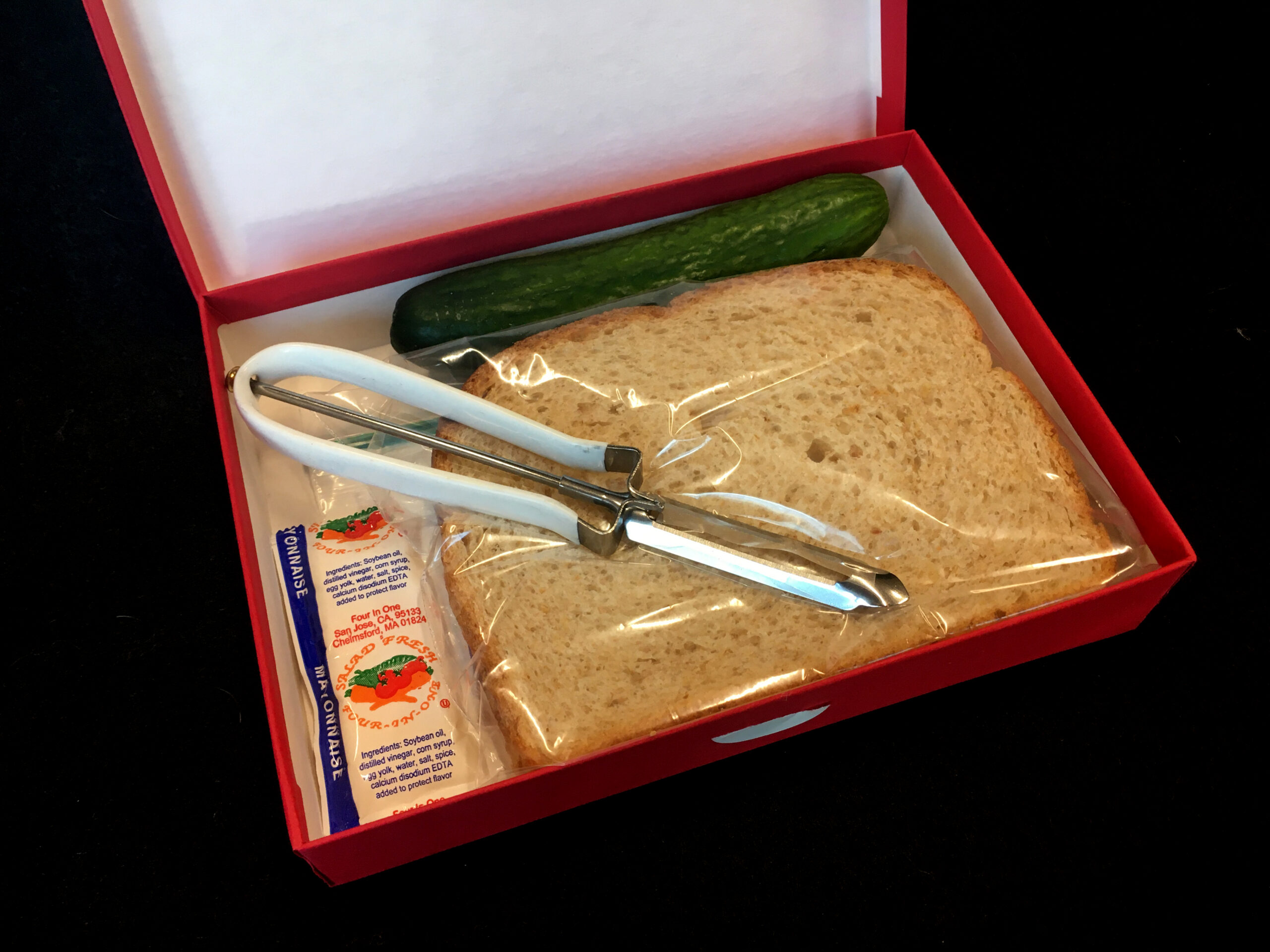 box contains paper, vegetable peeler, bread, cucumber, and mayonnaise packet